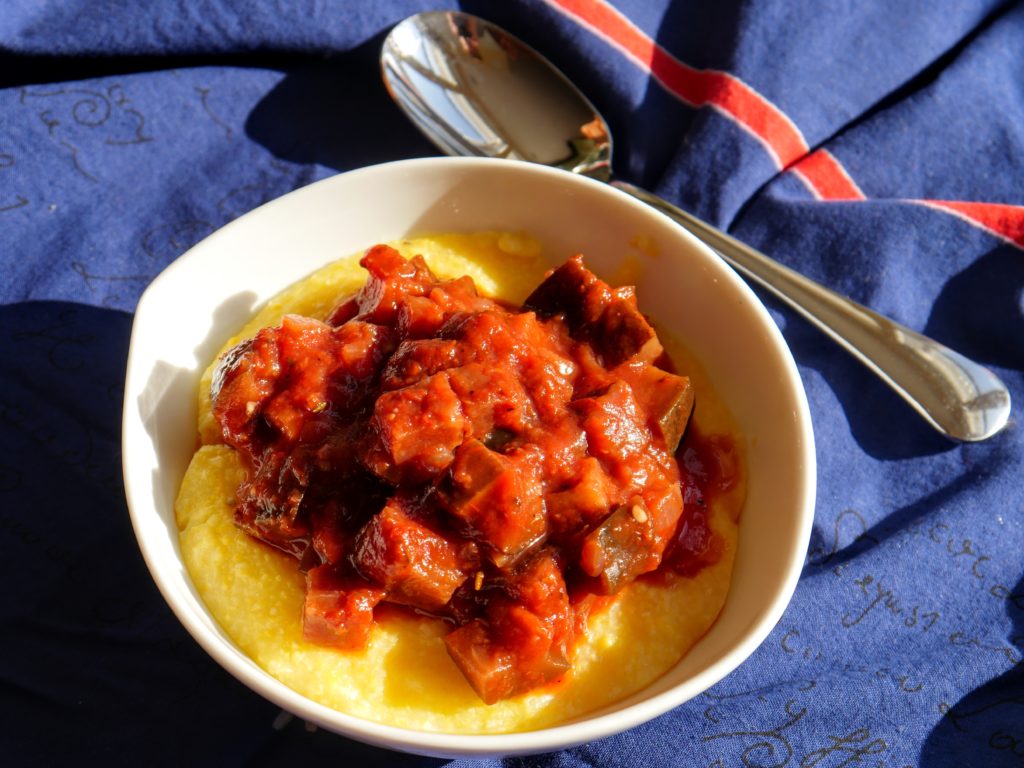 A bowl of polenta topped with eggplant-tomato ragout sits on a blue and red cloth.