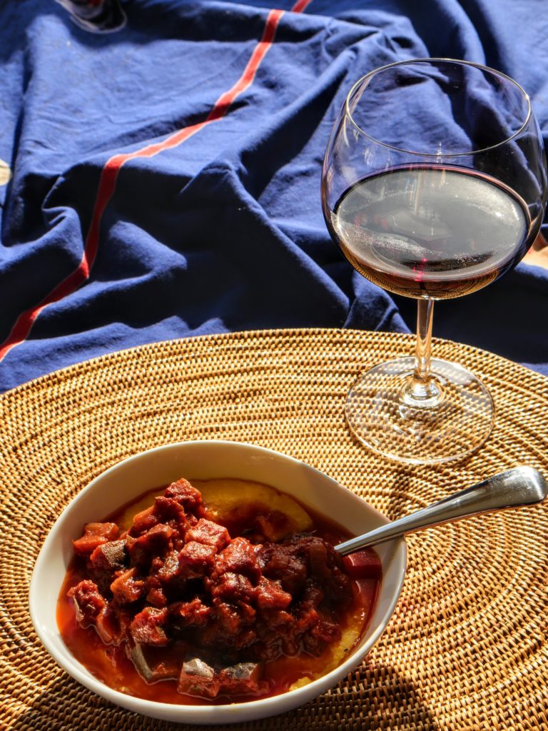 A bowl of polenta topped with eggplant-tomato ragout and a glass of red wine sit on a woven straw mat.