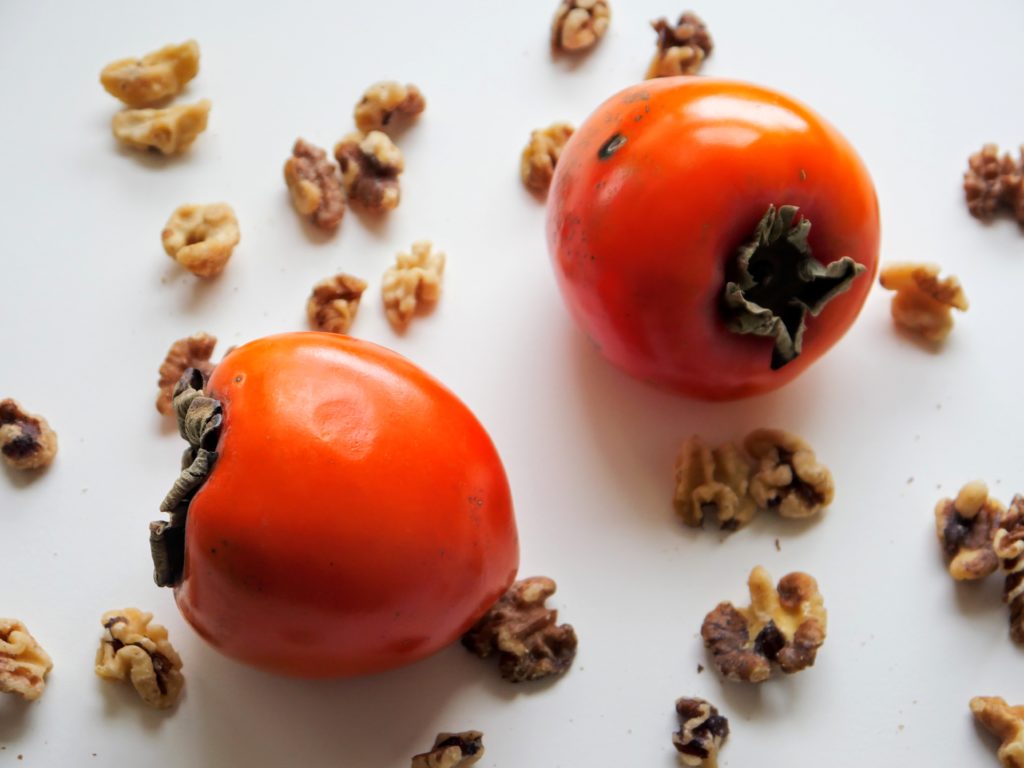 Two ripe persimmons sit on a white surface, surrounded by scattered walnuts.