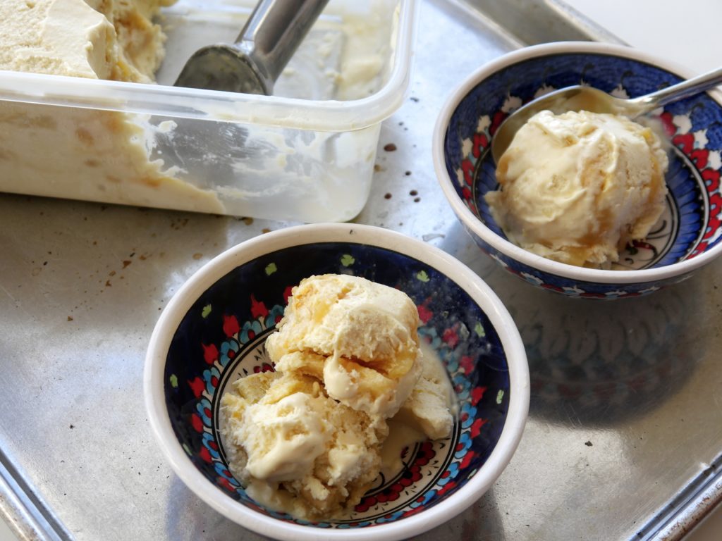 Two blue-red-and-white patterned bowls containing scoops of pale yellow ice cream sit on a metal surface next to a clear plastic tub of ice cream.