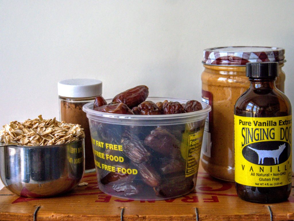 Oats in a measuring cup, a glass jar of cinnamon and one of peanut butter, dates, and a bottle of vanilla sit on top of a wooden surface in front of a white wall.