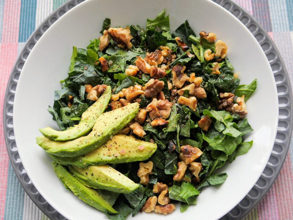 A white bowl - full of kale, arugula, walnuts, and sliced avocado - rests on a grey plate, both on a pastel-plaid placemat.