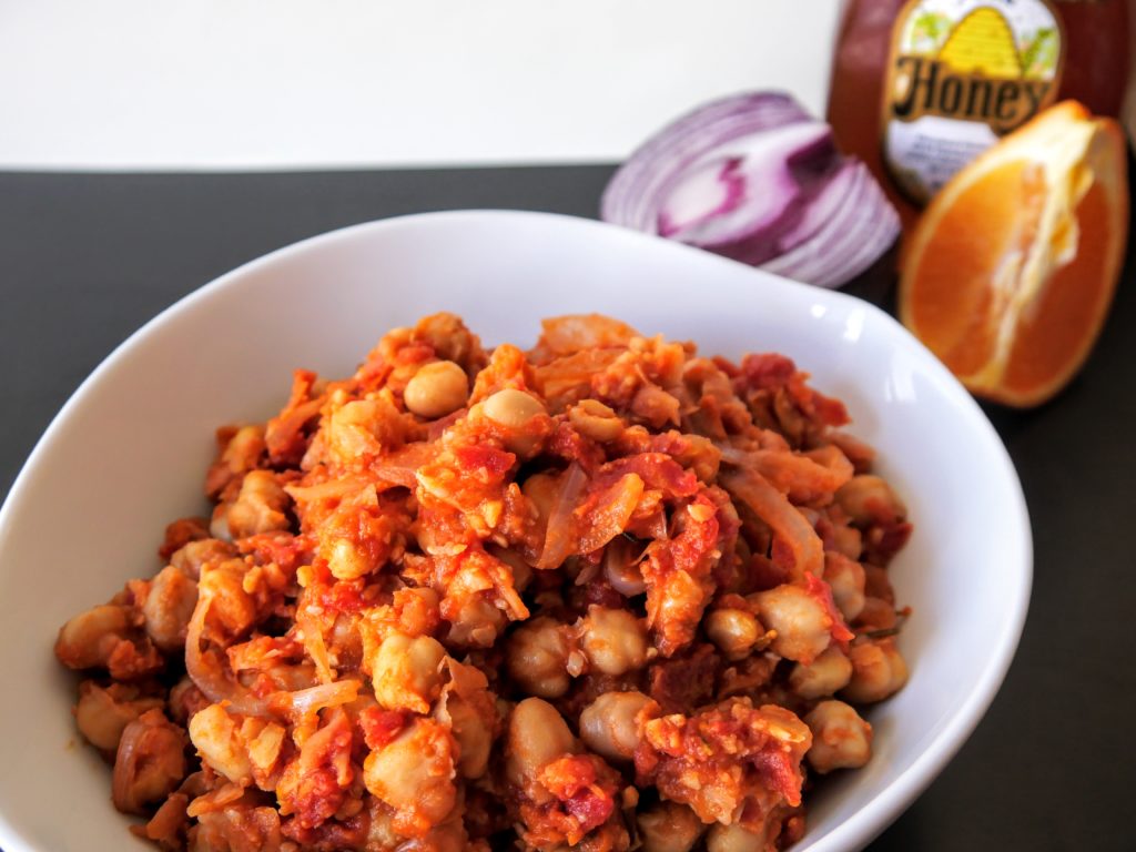 Chickpeas, cooked with tomatoes and onions, sit in a white bowl on a black surface. A halved red onion,  quartered orange, and jar of honey lay beyond the bowl.