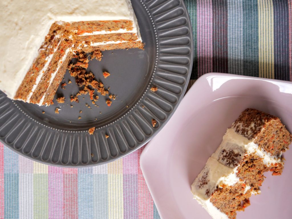 A three-layer carrot cake coated with cream cheese frosting sits on a gray plate, next to a slice of the cake that rests on a pink plate. Both sit on a pastel plaid cloth.