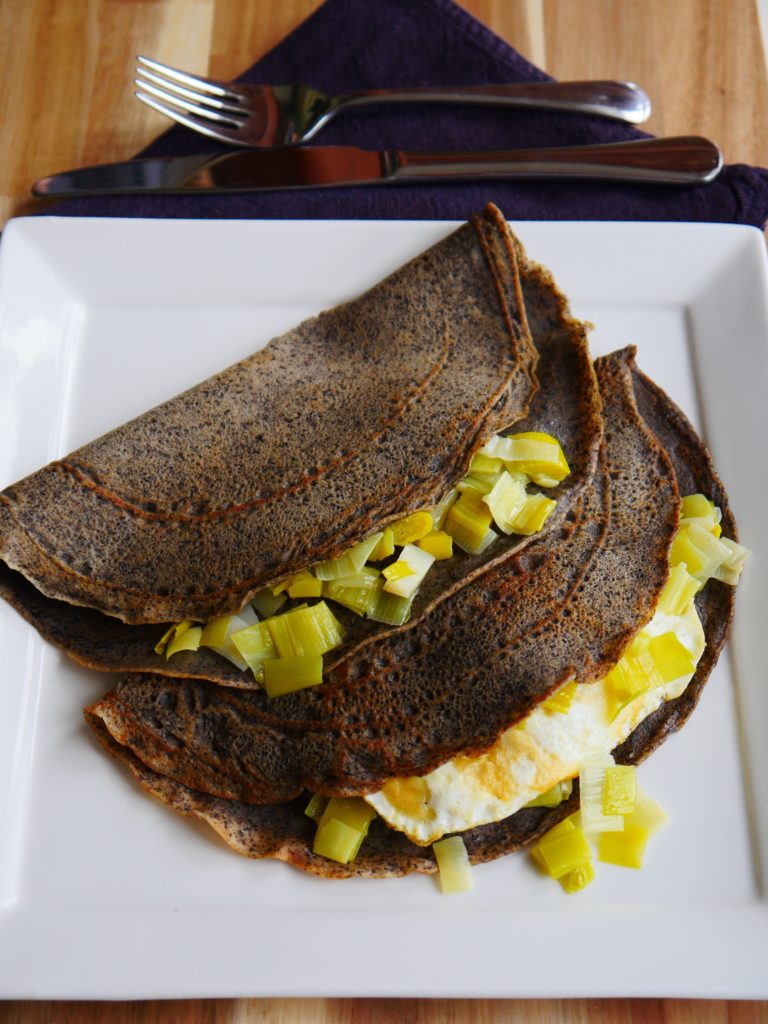 Two brown buckwheat crepes stuffed with leeks and one with a fried egg rest on a white plate, itself atop a wooden surface. A knife, fork, and dark purple napkin sit at the top edge of the shot.