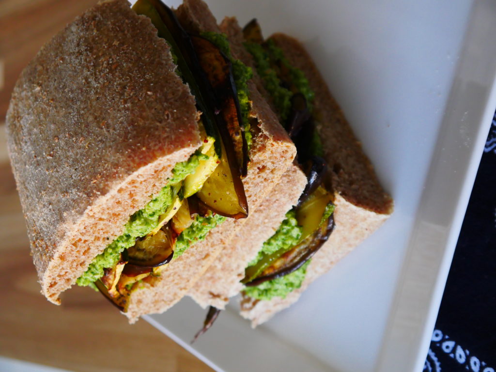 Two sandwiches - roasted vegetables and cilantro-jalapeno pesto on whole-wheat ciabatta - are stacked on top of each other, resting on a white plate.