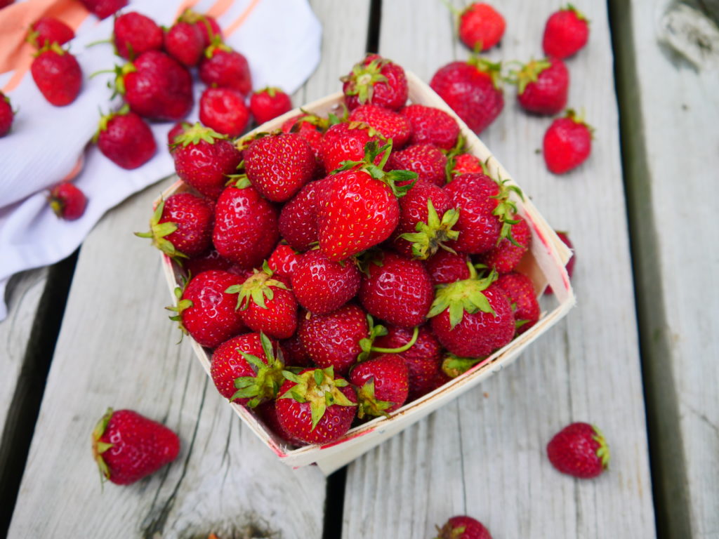 A small box of strawberries sits on a wood plank deck. Strawberries are spilling out of the box, onto the deck and a piece of white cloth in the upper-left corner of the photo.