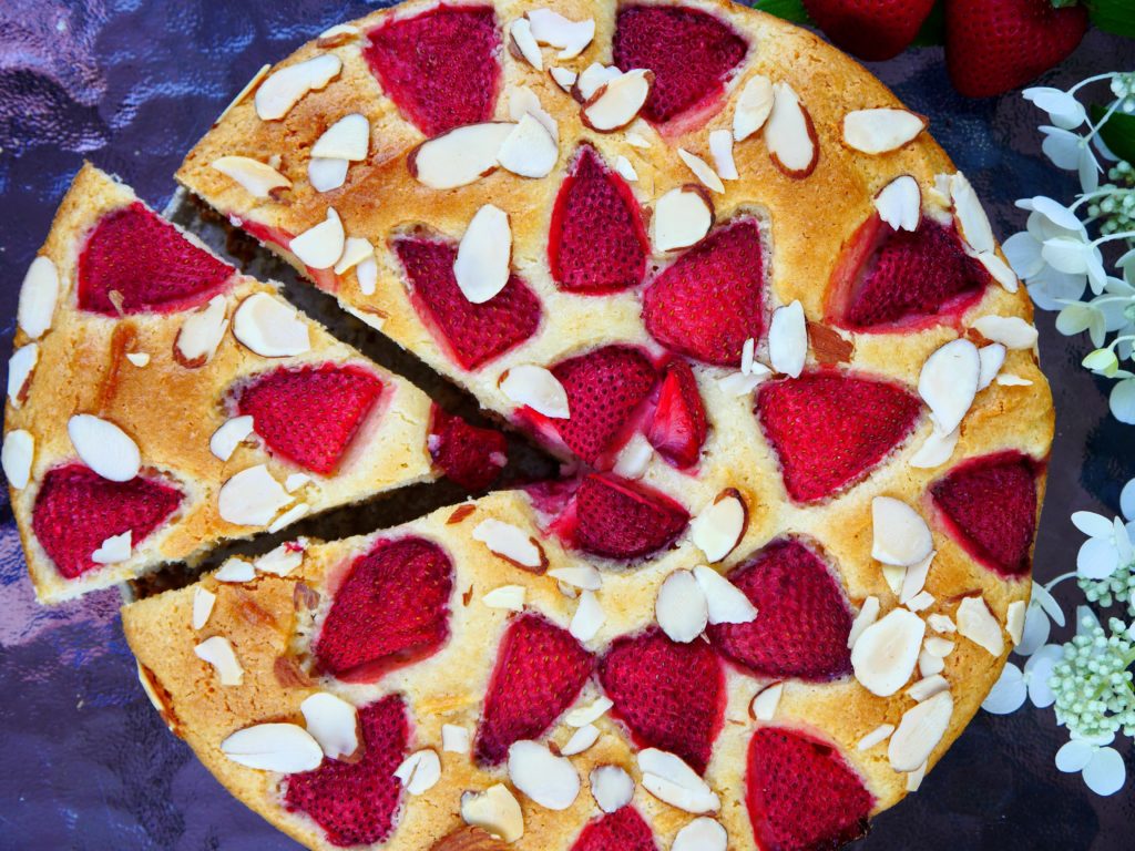 A slice of cake has been cut and is being lifted from golden brown cake, studded with ruby-red strawberries and slivered almonds.