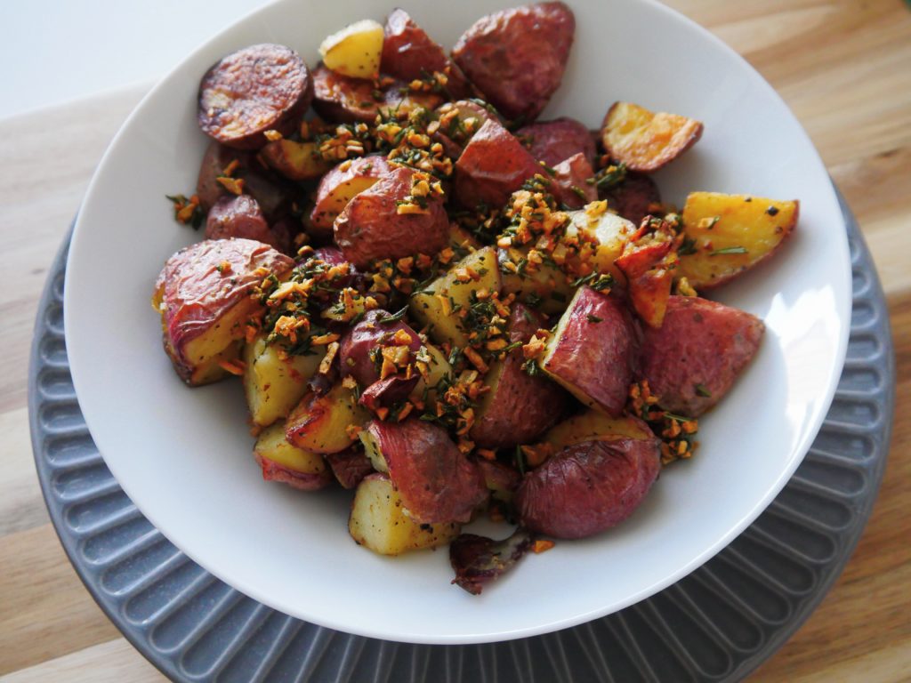 Roasted red potatoes, decorated with sauteed garlic and rosemary, sit in a white bowl on a wooden surface.