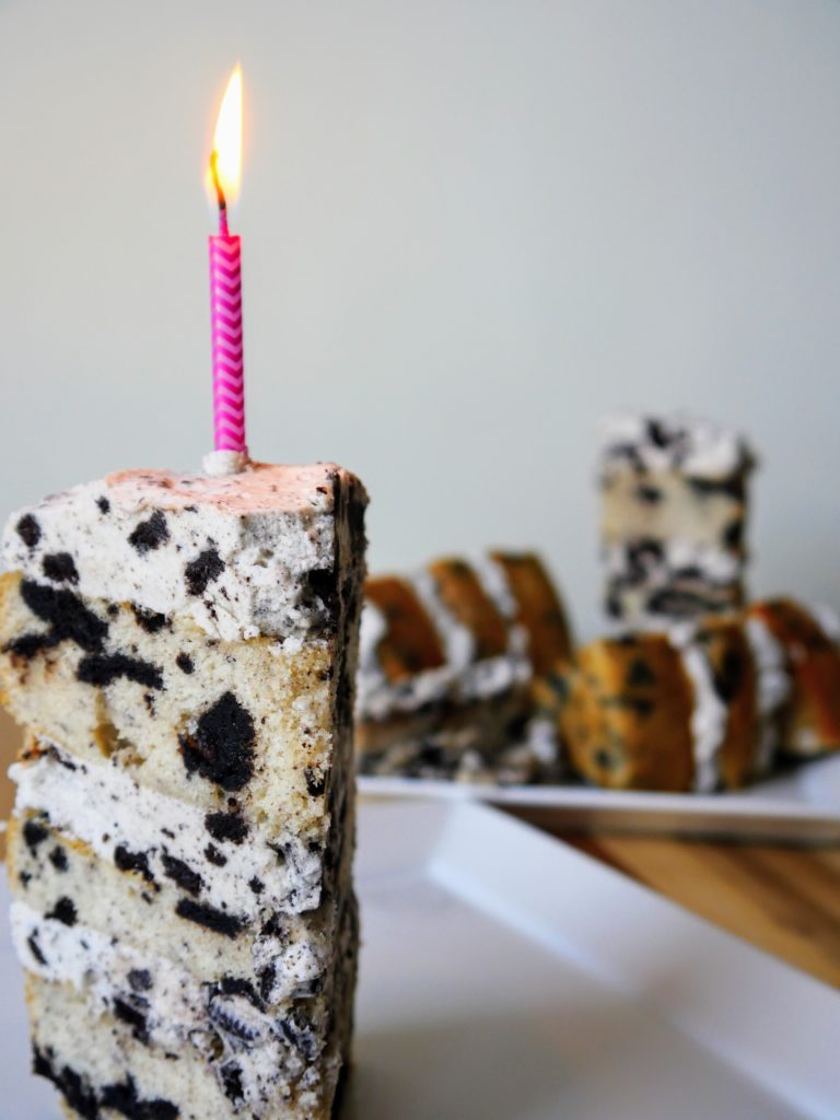 A slice of Oreo-studded white cake, layered with Oreo-studded vanilla frosting and topped with a pink birthday candle, sits on a white plate in the foreground. Behind it to the left, slices of cake that weren't as pretty as Sarah wanted rest on another white plate.