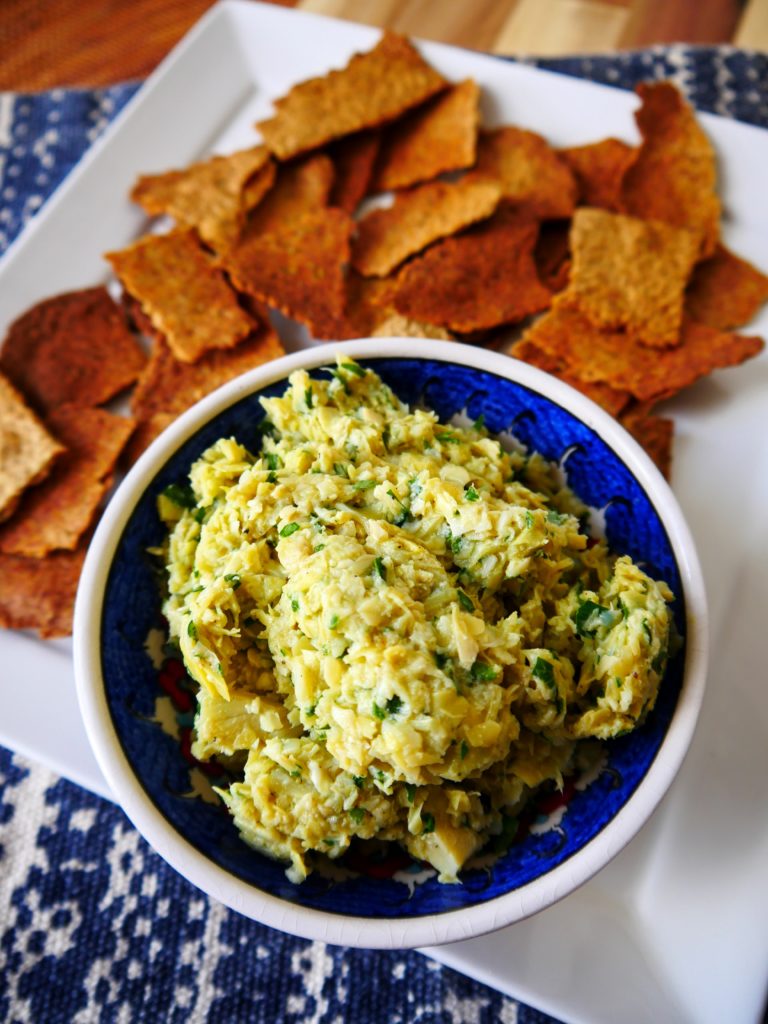 A blue bowl full of artichoke pâté sits on a white plate. The bowl is surrounded by toasty brown crackers.