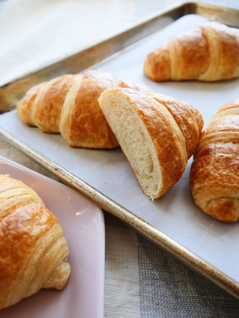 Several golden brown croissants rest on a parchment-paper lined baking sheet. Next to the sheet, another croissant sits on a pale pink plate.