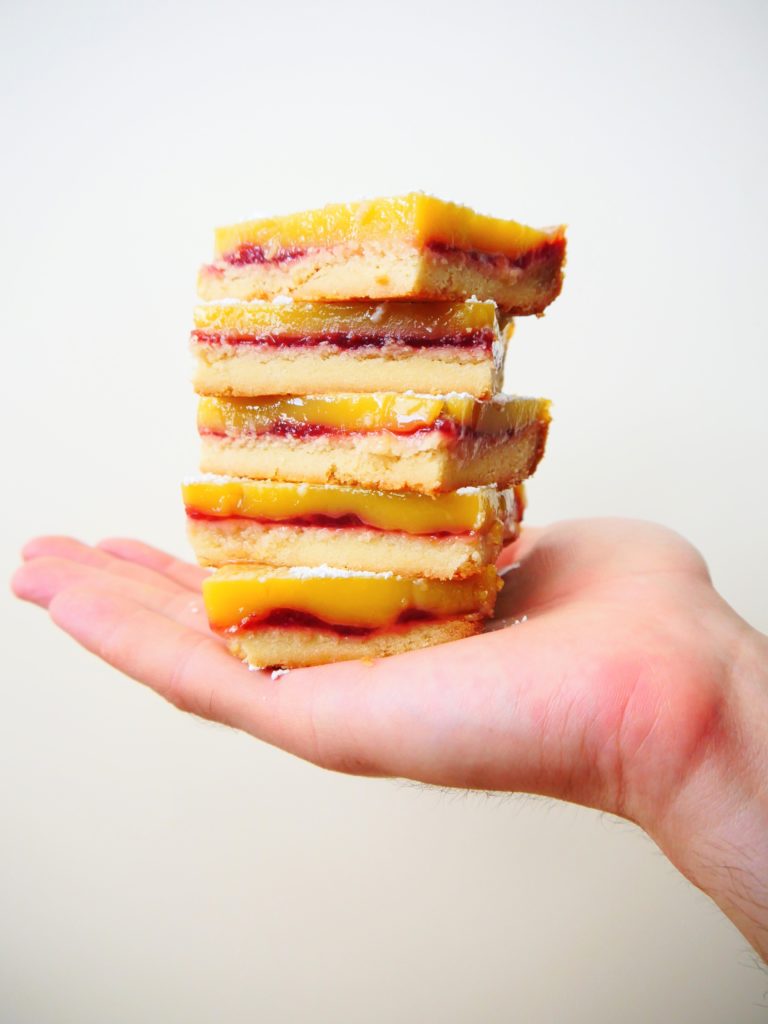 Five lemon raspberry bars (shortbread crust, raspberry jam, and lemon curd) sit stacked on Sarah's Chief Recipe Taster's hand.